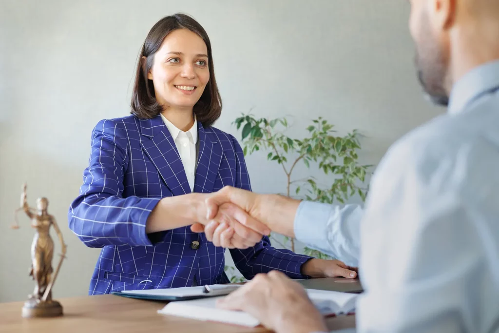 Image of a Caucasian women who is a licensed Notary shaking hands with a bearded man who is a lawyer, the woman has a blue suit jacket on with white lines forming squares, she has blue eyes, and is smiling as they finalize the deal on starting a notary business.  