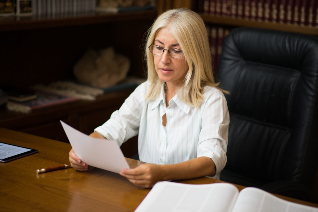 Blonde haired woman with classes and a white and blue stripe shirt reading a document on Notary Liability