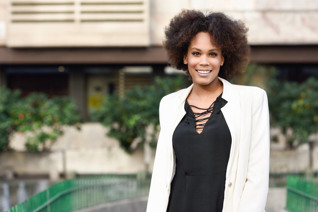 Young notary signing agent with afro hairstyle smiling in urban background. Mixed girl wearing white jacket and black dress.