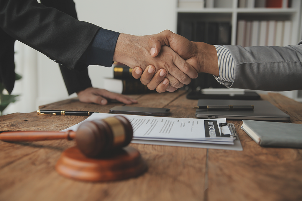two men in suits shaking hands over a table. The table has documents, pens, a cell phone, laptop, and gavel. Notary status in Massachusetts.